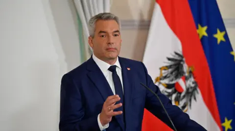 Karl Nehammer, portant un costume bleu foncé et une cravate, se tenant devant un microphone, avec les drapeaux autrichien et de l'UE derrière lui.