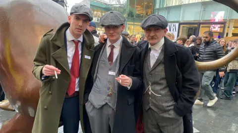 Trois hommes en casquettes grises et manteaux, deux d'entre eux tenant des cigarettes.