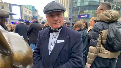 Un homme portant une casquette noire et un manteau noir avec un nœud papillon noir sur une chemise blanche.