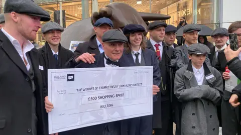 Des hommes et des femmes habillés en casquettes et manteaux devant une statue de taureau en bronze. Un homme tient un gros chèque blanc indiquant un bon d'achat de 500 euros.