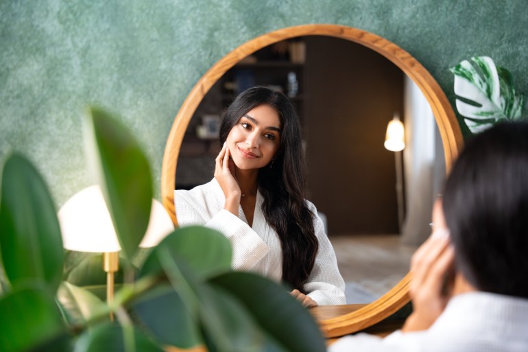 Soins capillaires et soins personnels avec une belle femme indienne regardant dans le miroir touchant ses longs cheveux sains assise à la coiffeuse.