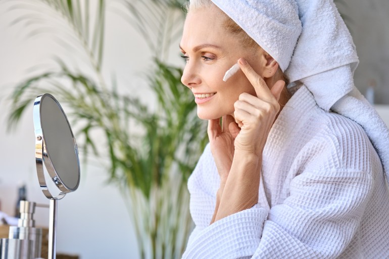 Femme blonde senior appliquant un traitement hydratant sur le visage dans la salle de bain, regardant dans le miroir.